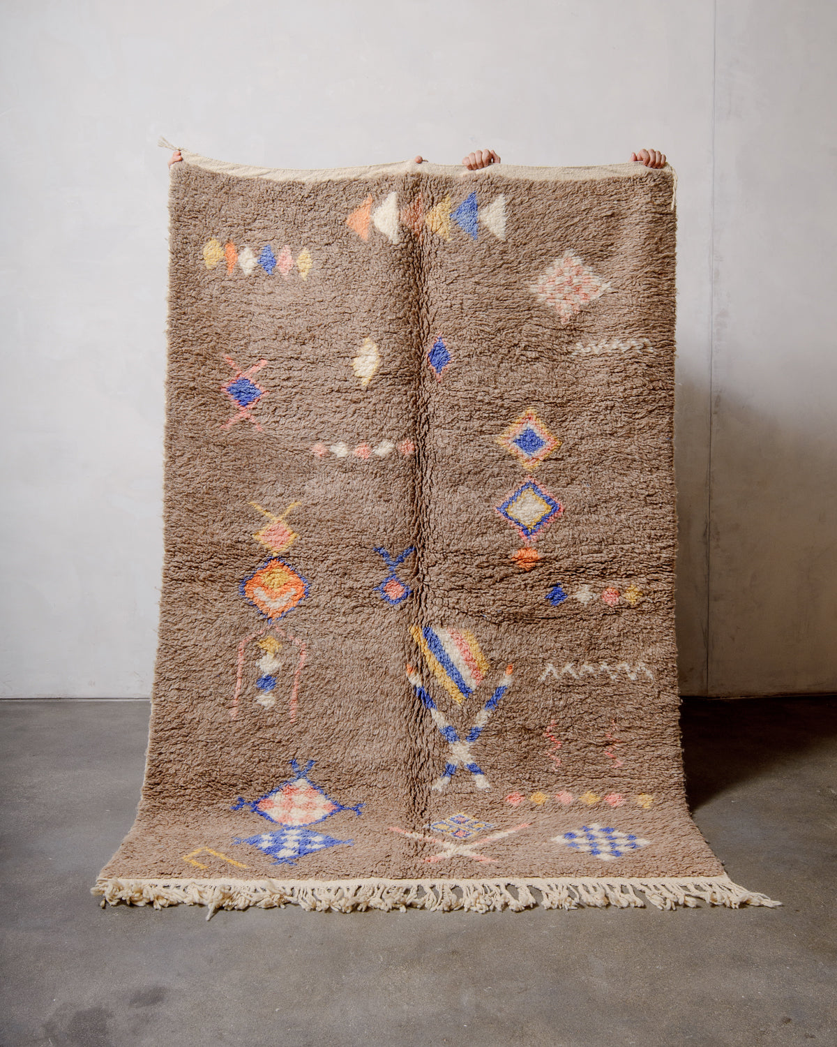 Berberlin Vintage Berber Rug made of fluffy cream wool, being held up against a white wall with a grey concrete floor.